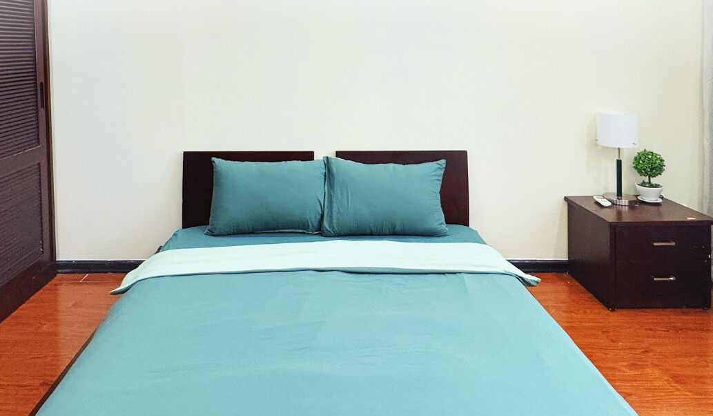 A neatly made bed with a teal bedspread and pillows, a bedside table with a lamp and a small plant, and a wooden floor.