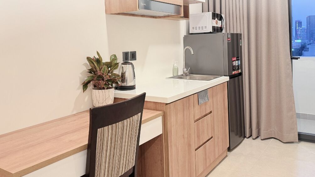Une cuisine compacte au design moderne, équipée d’un four à micro-ondes encastré, d’un réfrigérateur et d’un évier. On y trouve une petite plante en pot sur le plan de travail et une chaise avec un coussin à rayures.