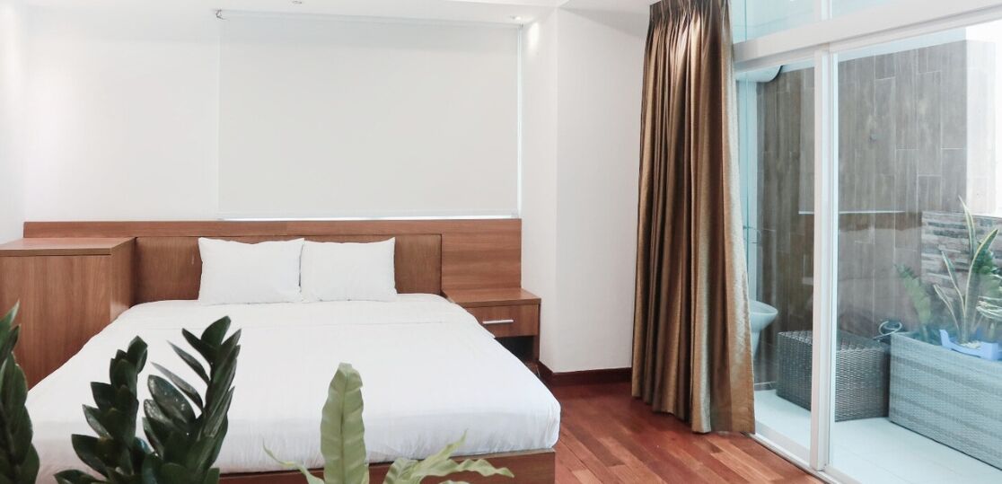 Una habitación de hotel moderna con una cama grande, cabecera de madera, una mesita de noche y una puerta corrediza de vidrio que da a un balcón con plantas.