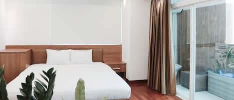 A modern hotel room with a large bed, wooden headboard, a nightstand, and a sliding glass door leading to a balcony with plants.