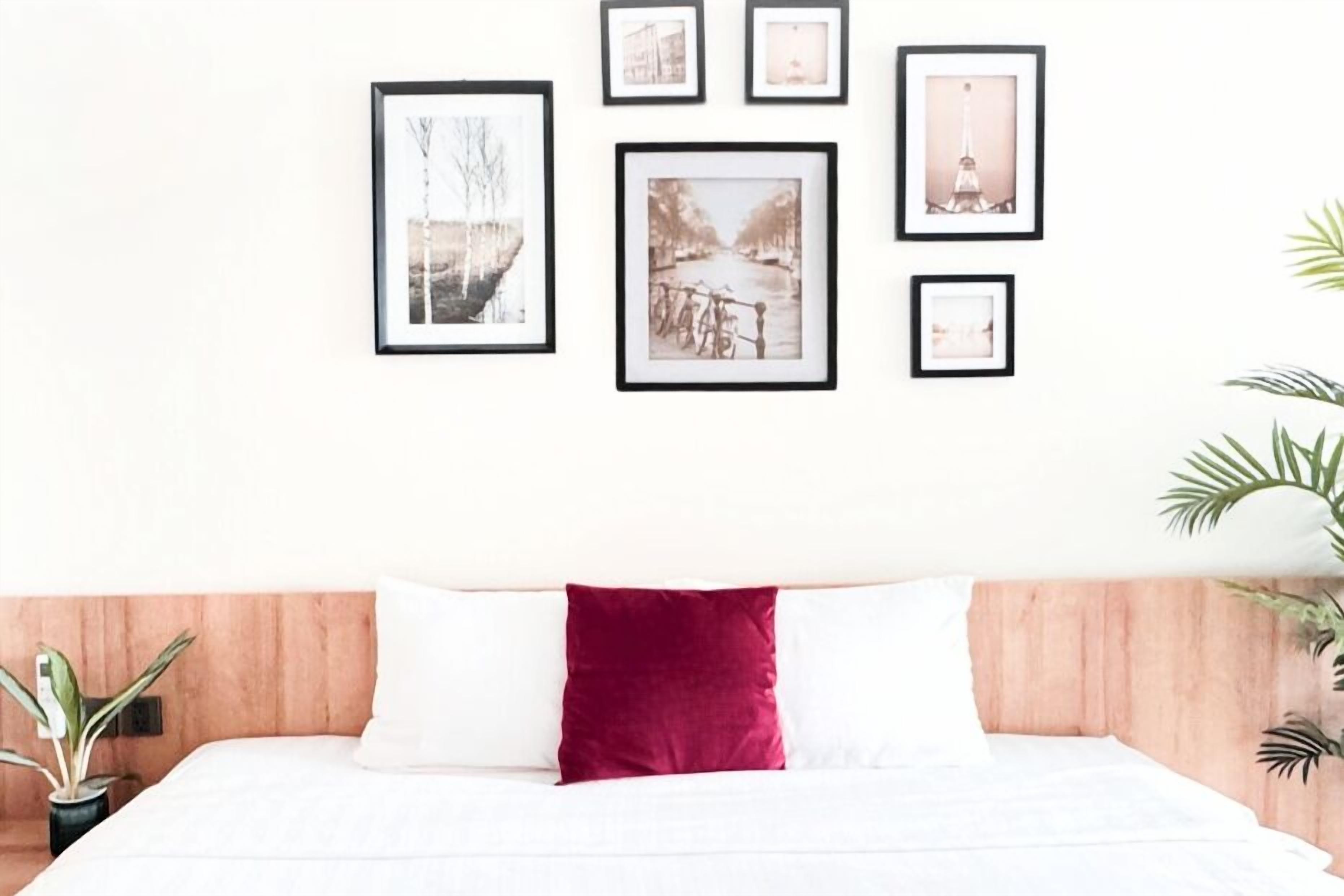 A neatly arranged bedroom with a wooden headboard, a white bedspread, and a red accent pillow. There are framed pictures on the wall, and potted plants on either side of the bed.