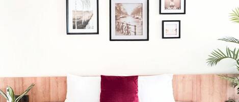 A neatly arranged bedroom with a wooden headboard, a white bedspread, and a red accent pillow. There are framed pictures on the wall, and potted plants on either side of the bed.