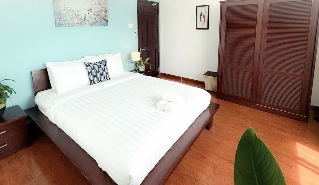 Una cama bien tendida con sábanas blancas y una almohada, una mesita de noche con una lámpara, una planta verde y un ventanal grande.