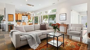 A spacious living area with a sofa, coffee table, and a chair. There is a kitchen in the background with wooden cabinets and a refrigerator.