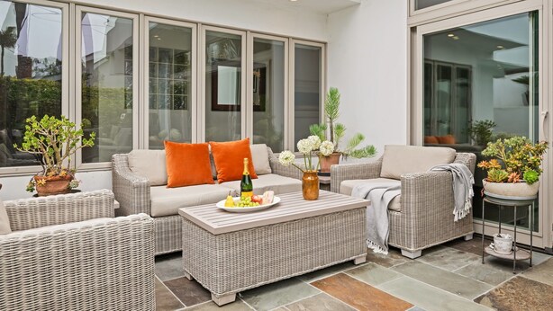 A sunlit patio area with wicker furniture, a coffee table with fruit and a bottle, and potted plants.