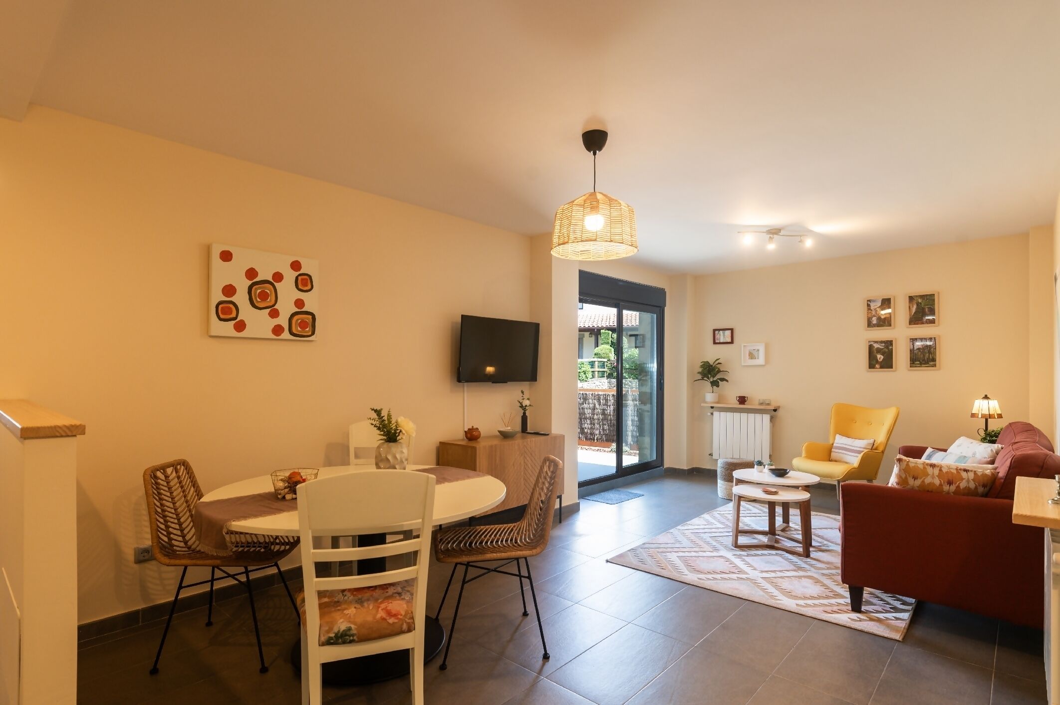A modern living room with a dining area, a TV mounted on the wall, and a large window providing natural light.