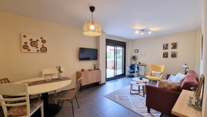 A modern living room with a dining area, a TV mounted on the wall, and a large window offering a view of the outdoors.