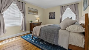 Un dormitorio con cama, cortinas, una mesita de noche, lámpara y cuadros en las paredes.