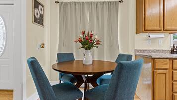 Een eetgedeelte met een ronde tafel, teal stoelen, een bloevas en een keuken met houten kasten.