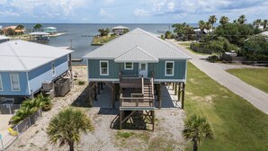 A beachfront property with a two-story house elevated on stilts, a covered deck, and a view of the ocean. - Bayfront Home with Beach Access (Gulf Shores)