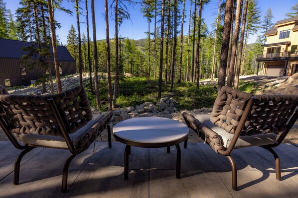A patio with two woven chairs and a round table, surrounded by a forested area and a building in the background. - Impressive modern ski and summer home. (Whitefish)
