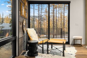 A room with a large window overlooking a forest, a wooden chair, a small stool, and a cowhide rug. - Impressive modern ski and summer home. (Whitefish)