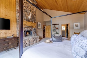 A bedroom with a stone fireplace, a bed, a TV, and a wooden ceiling.