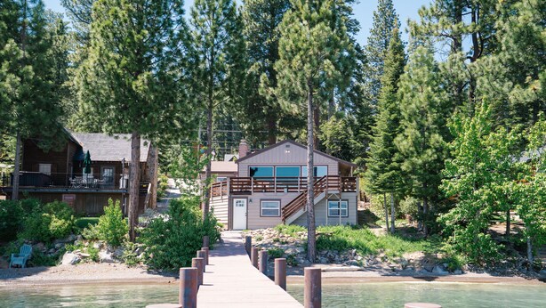A wooden dock leading to a cabin with a large window and a deck, surrounded by dense trees and a clear water body.