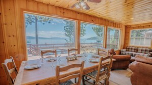 A wooden dining area with a table set for six, large windows with a view of a lake and dock, and a cozy living area with a sofa and armchairs.
