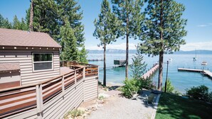 A cabin with a deck overlooking a lake with boats and mountains in the background.