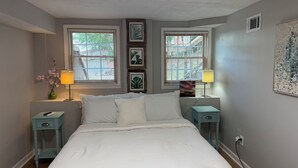 A bedroom with a bed, two bedside tables, a lamp, a picture frame, and a window with blinds. - Pleasantly Charming 2-bedroom duplex in  Wynnton Village Historic District. (Columbus)