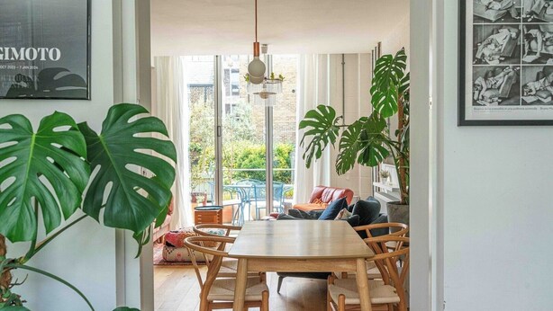 Een eethoek met een houten tafel en stoelen, grote groene planten en uitzicht op buiten.