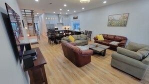 A spacious living area with a wooden floor, a brown leather sofa, a grey sofa, and a brown leather armchair. There is a dining table with chairs and a kitchen area in the background.