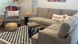 Un salon comprenant un canapé d’angle gris, un pouf rond, un tapis à motifs noir et blanc, une lampe sur pied, une petite table d’appoint et une étagère murale agrémentée d’objets décoratifs.
