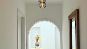 Un couloir avec une porte en arc donnant sur une pièce, un vase contenant des fleurs séchées posé sur un piédestal, et un chapeau sur une table.
