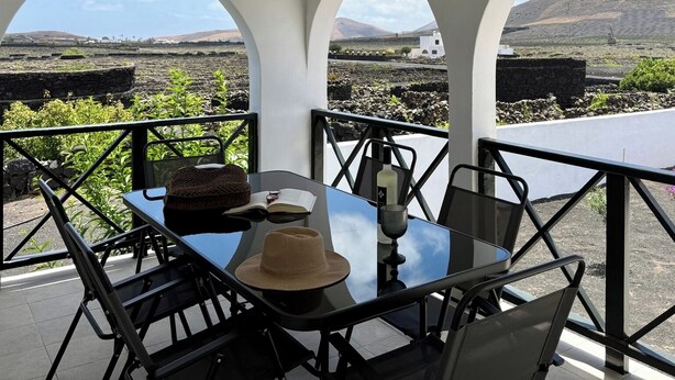 Une table dressée avec un chapeau, un livre et une bouteille, donnant sur un paysage pittoresque avec une montagne et des bâtiments blancs.