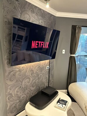 A modern hotel room with a large flat-screen TV mounted on the wall displaying the Netflix logo, a small bedside table with a remote control, and a window with curtains. - Stylish Family 1BR + Cozy Bunks for 5 with Games (Darlinghurst)