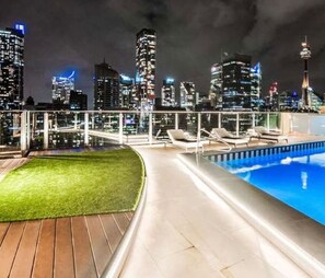 Une piscine sur le toit offrant une vue panoramique sur la ville, des chaises longues et un espace verdoyant.