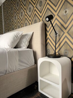 A bedroom with a bed, pillows, a nightstand with a lamp, and a patterned wallpaper.