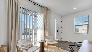 Ein modernes Zimmer mit Holzboden, einem Esstisch, zwei Stühlen, einem kleinen runden Tisch und einem großen Fenster mit Jalousien.