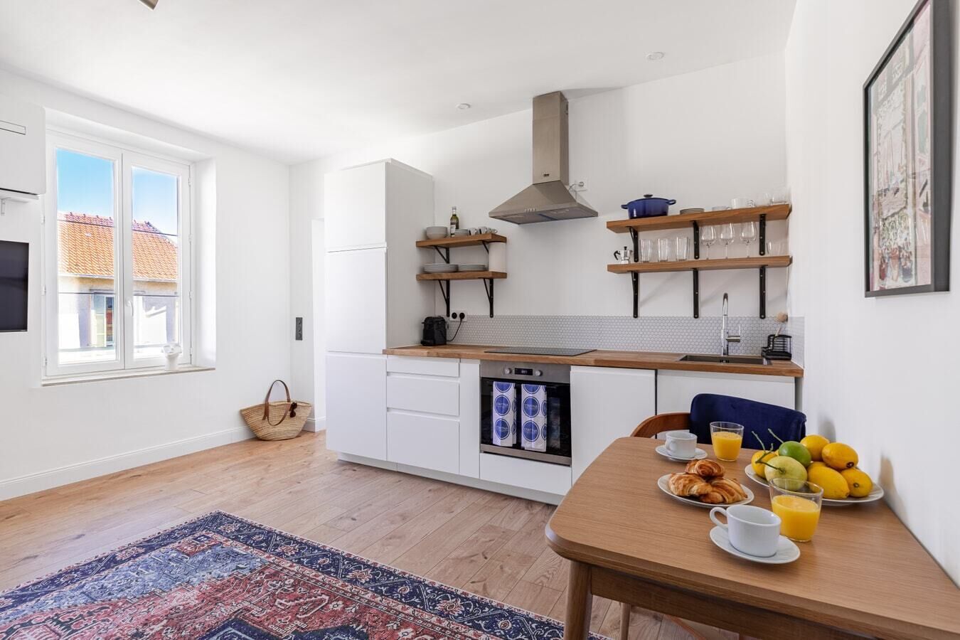 A modern kitchen with white cabinets, a built-in oven, and a large window. There is a dining table with breakfast items and a rug on the floor.