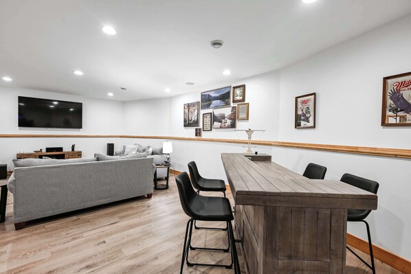 Una sala de estar moderna con un sofá gris, una mesa de comedor de madera con taburetes altos y un televisor de pantalla plana montado en la pared.
