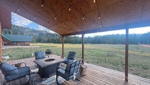 A covered outdoor seating area with a fire pit, chairs, and a view of a forested landscape.
