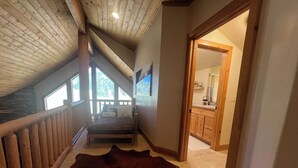 A bedroom with a large window, a wooden ceiling, a stone wall, a bed, a rug, and a bathroom visible through an open door.