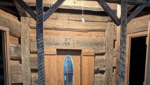 Interior rústico de una cabaña con paredes de madera, un ventanal central y una pequeña mesa con una botella y una taza.