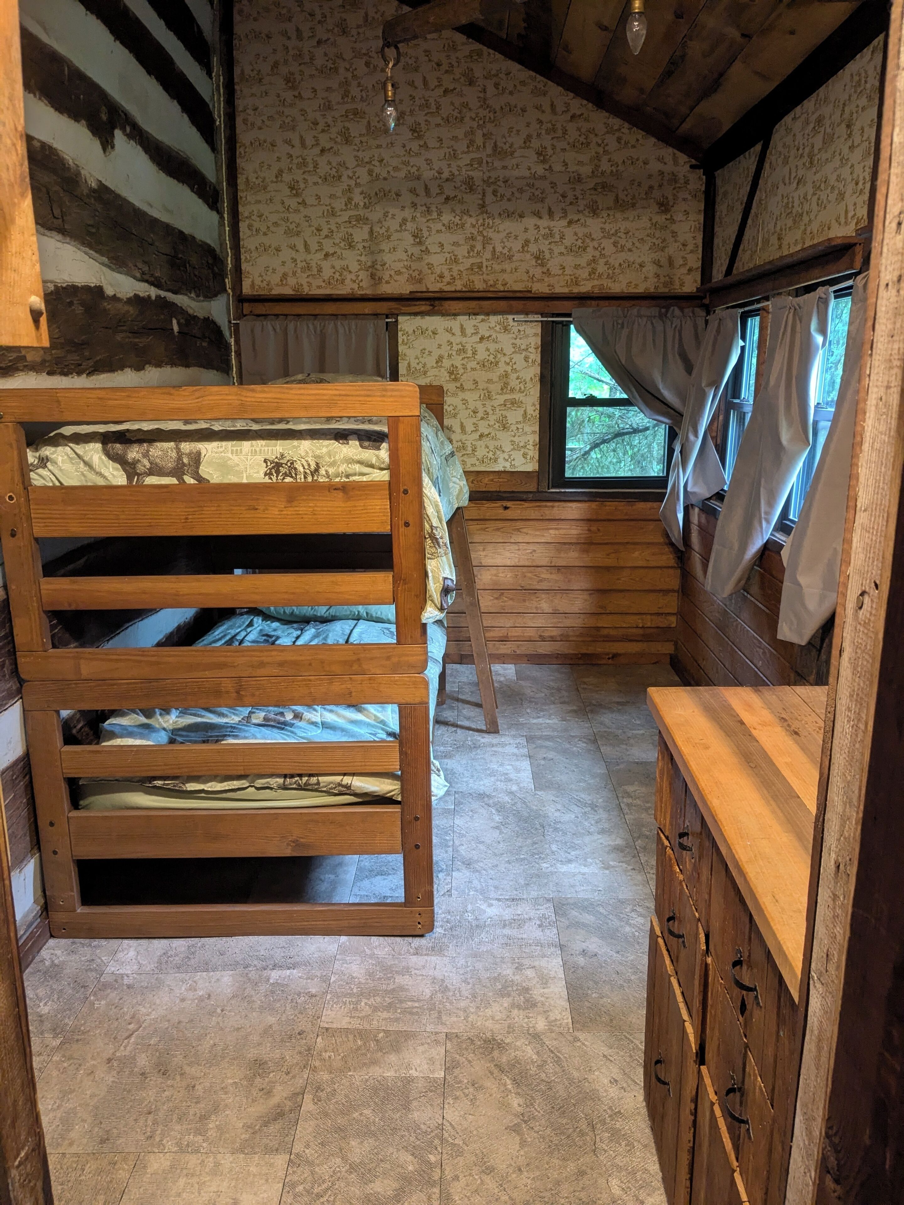 Ein rustikaler Holzraum mit zwei Etagenbetten, einem kleinen Holzschrank und einem Fenster mit Vorhängen.