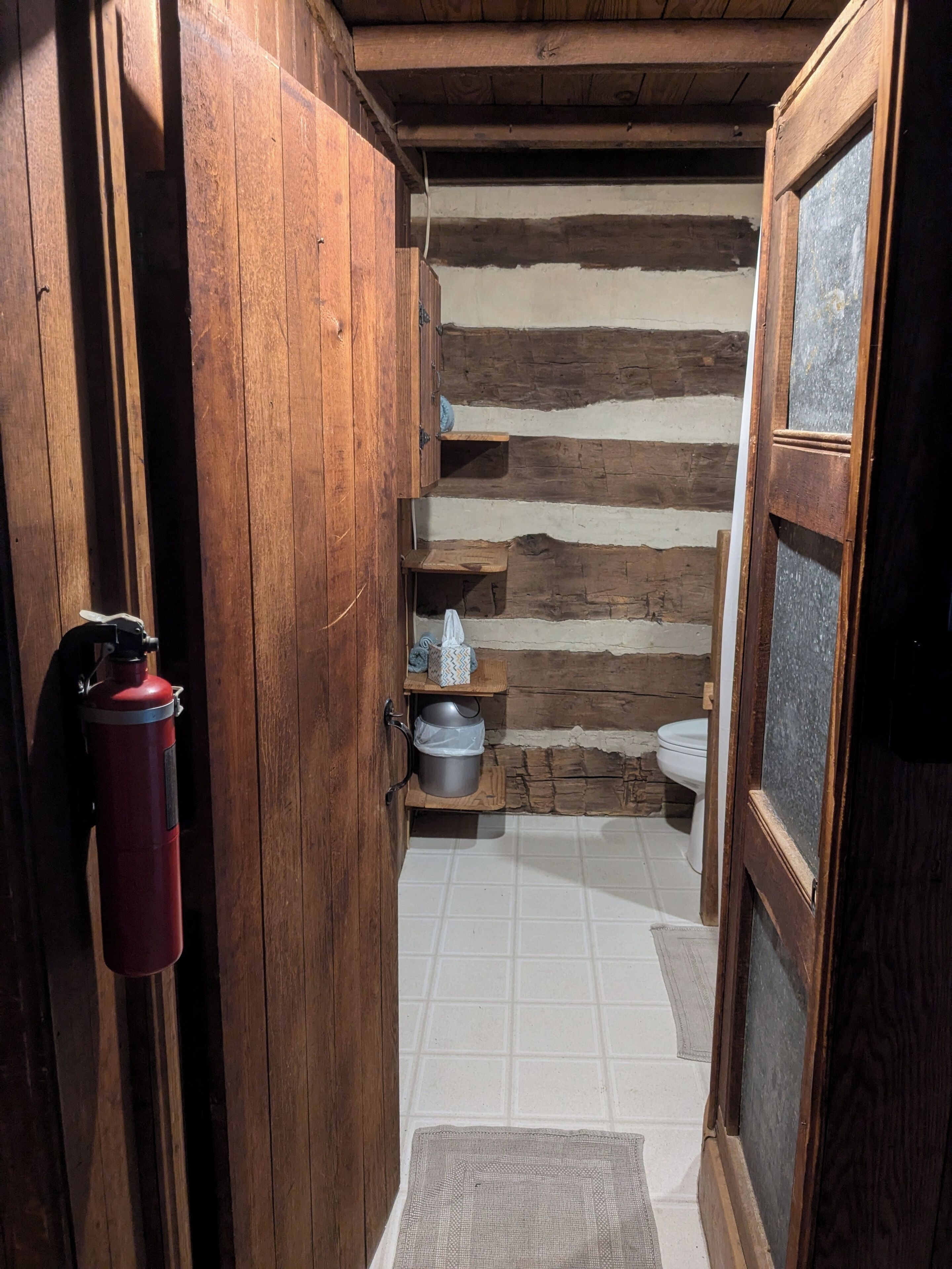 A narrow wooden hallway leading to a rustic bathroom with a wooden wall, a tiled floor, and a wooden door.