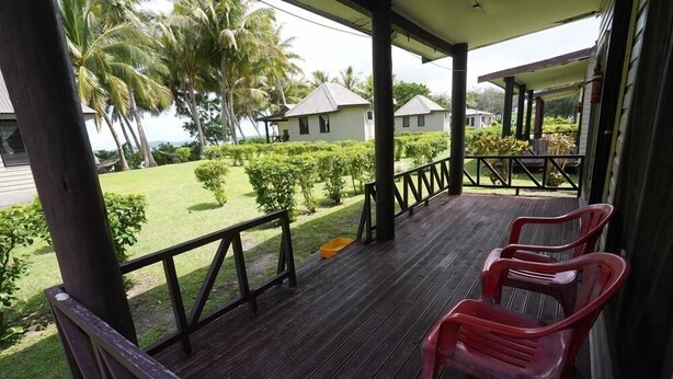 A wooden deck with a railing, two red plastic chairs, and a view of a well-maintained lawn and palm trees.