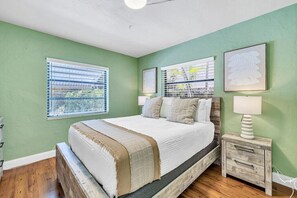Quarto com cama, duas janelas com persianas, mesa de cabeceira com abajur e obras de arte nas paredes.