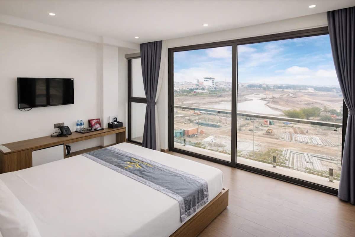 Hotelkamer met een groot bed, een televisie aan de muur, een bureau met telefoon en waterfles, en uitzicht op een bouwplaats door grote ramen.