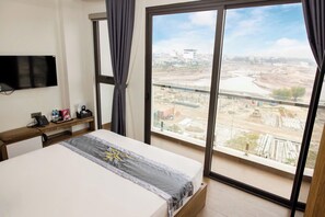 Quarto de hotel com cama, televisão na parede, mesa de cabeceira com telefone e garrafa de água, e uma janela grande com vista para uma obra.