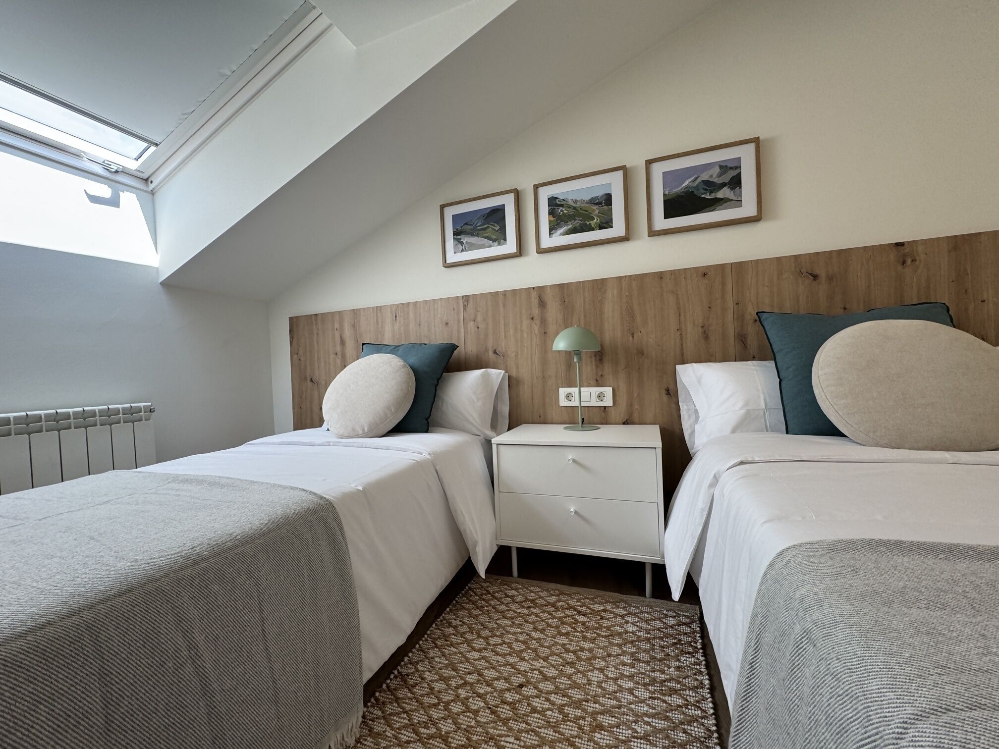 Un dormitorio con dos camas, cabeceros de madera, una mesita de noche con lámpara y tres cuadros enmarcados en la pared.