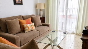 Una sala de estar con un sofá, una mesa de centro de vidrio, una lámpara y vistas al exterior a través de una ventana.
