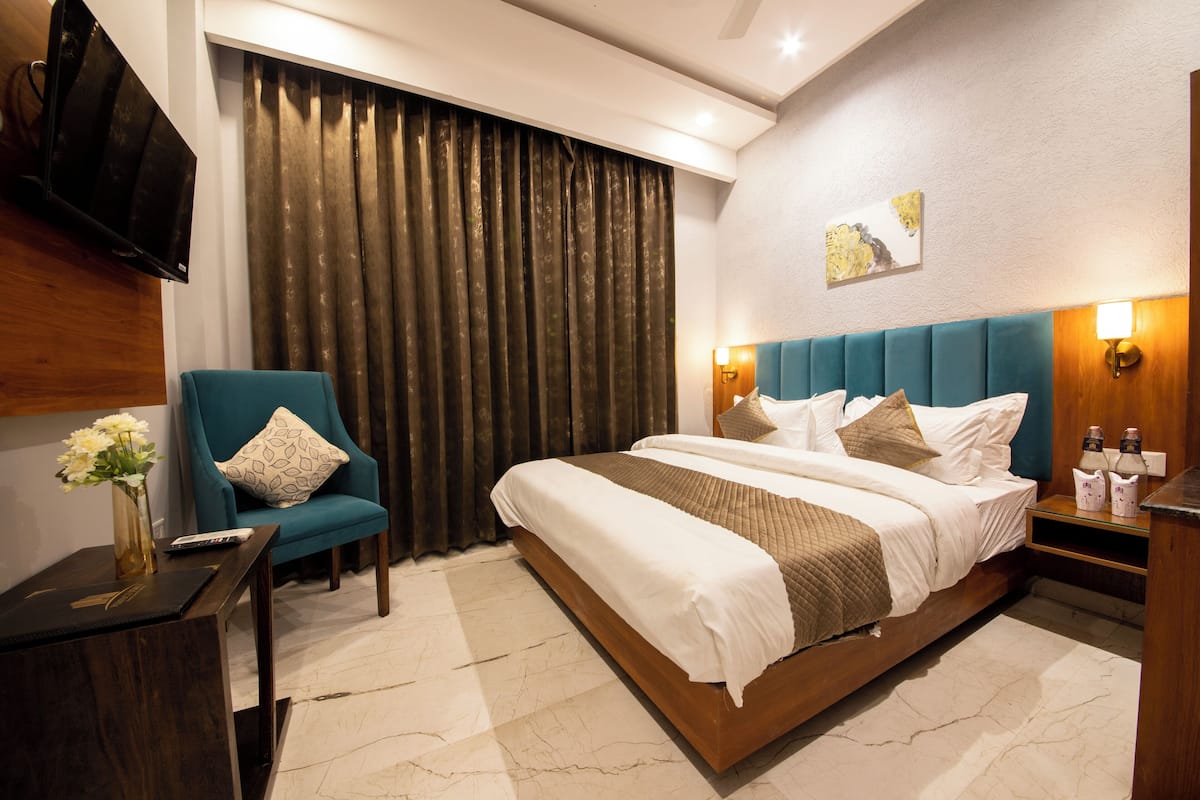 Ein Hotelzimmer mit einem großen Bett, einem Sessel, einem Nachttisch, einem an der Wand montierten Fernseher und einem Vorhang.