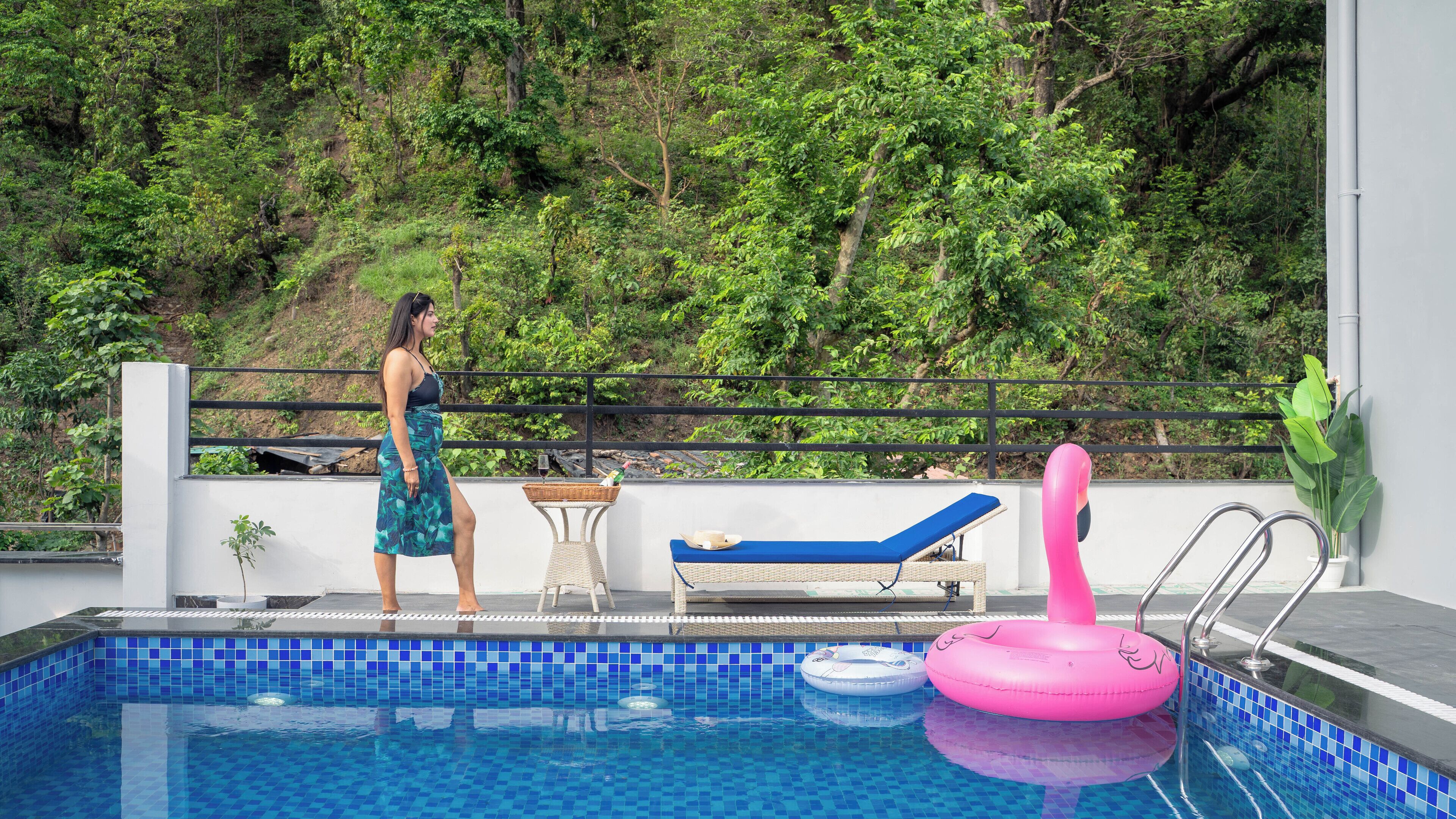 A swimming pool with a tiled blue border, a lounge chair, a pink inflatable float, and a view of a forested hillside.