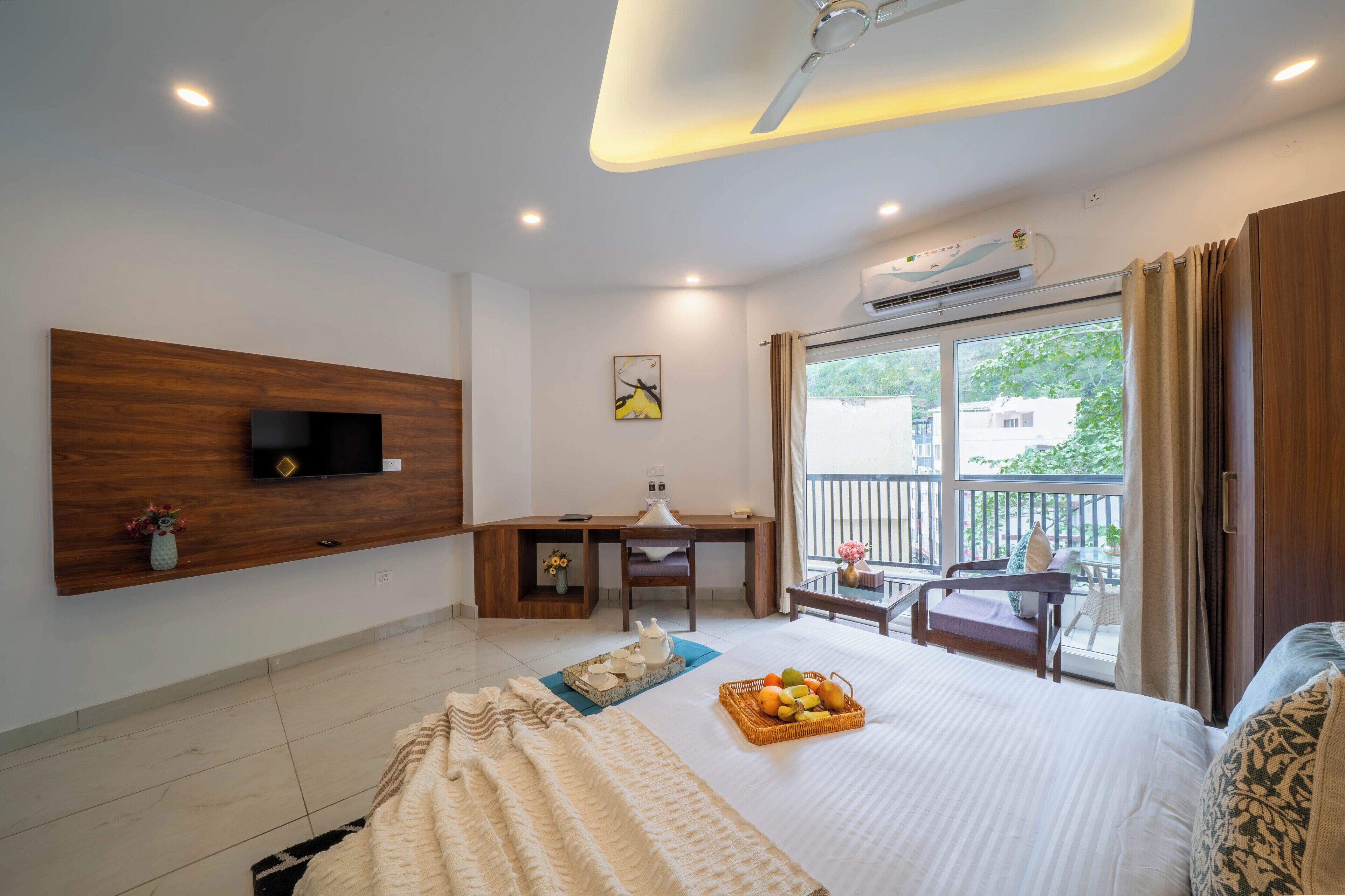 A modern hotel room with a large bed, a desk with a chair, a TV mounted on the wall, and a balcony with a view of the outside.