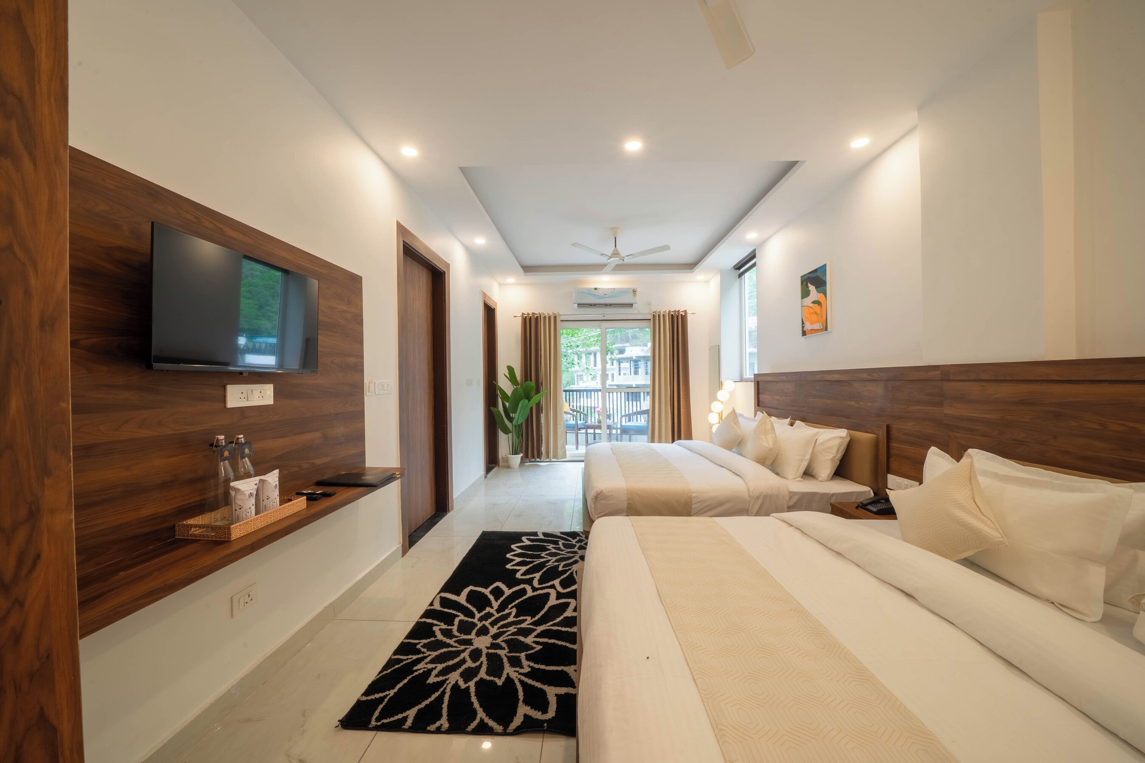 Una habitación de hotel con un televisor montado en la pared, una cama con ropa blanca, una mesita de noche y un amplio ventanal con cortinas.