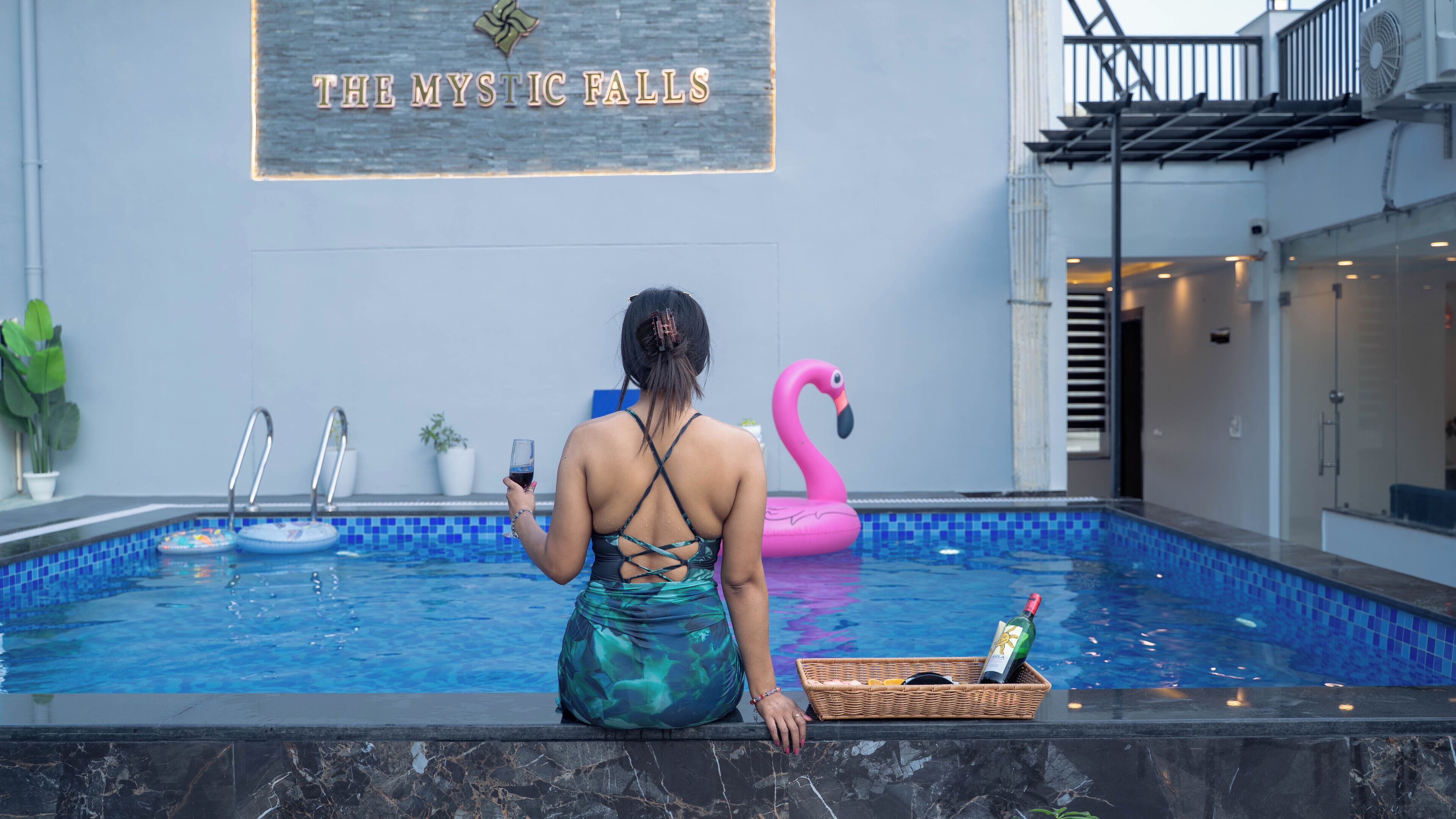 Une personne est assise au bord d’une piscine intérieure, avec un flotteur en forme de flamant rose, une bouteille de vin et un panier à proximité.