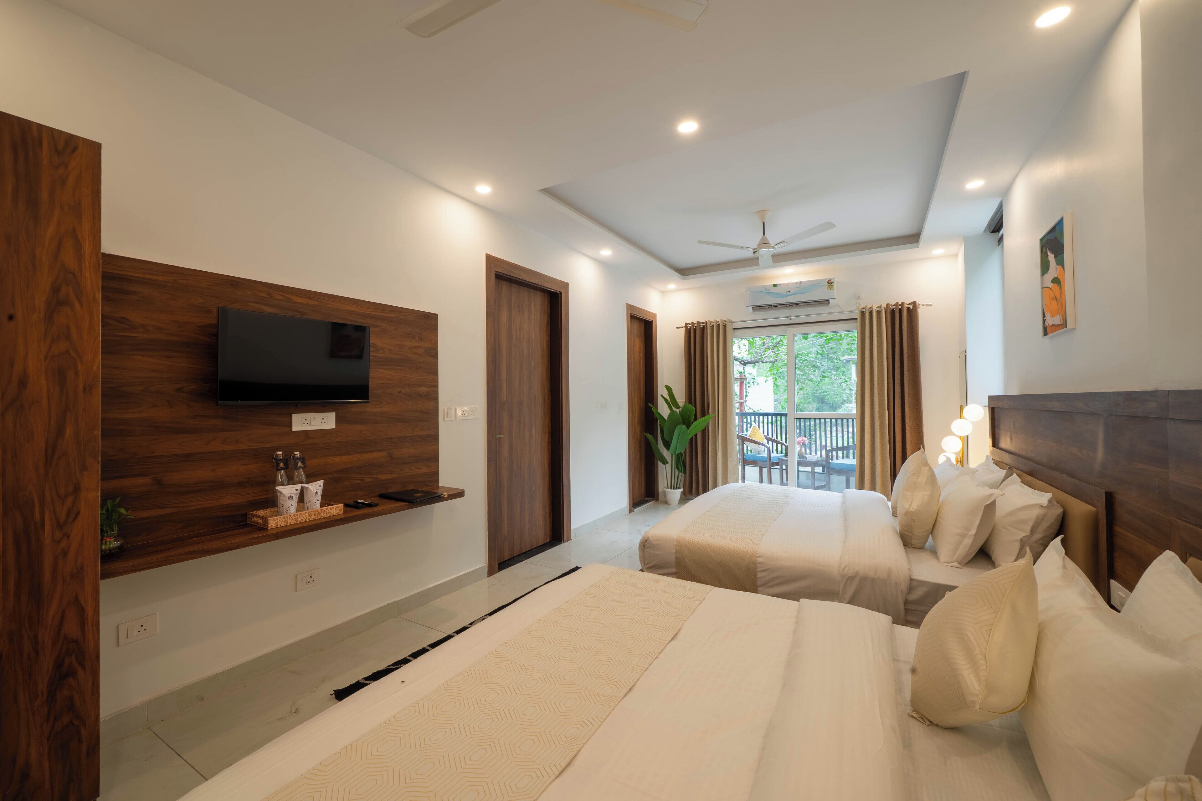 Habitación de hotel con dos camas, un televisor montado en un panel de madera, un balcón con vistas y un ventilador de techo.
