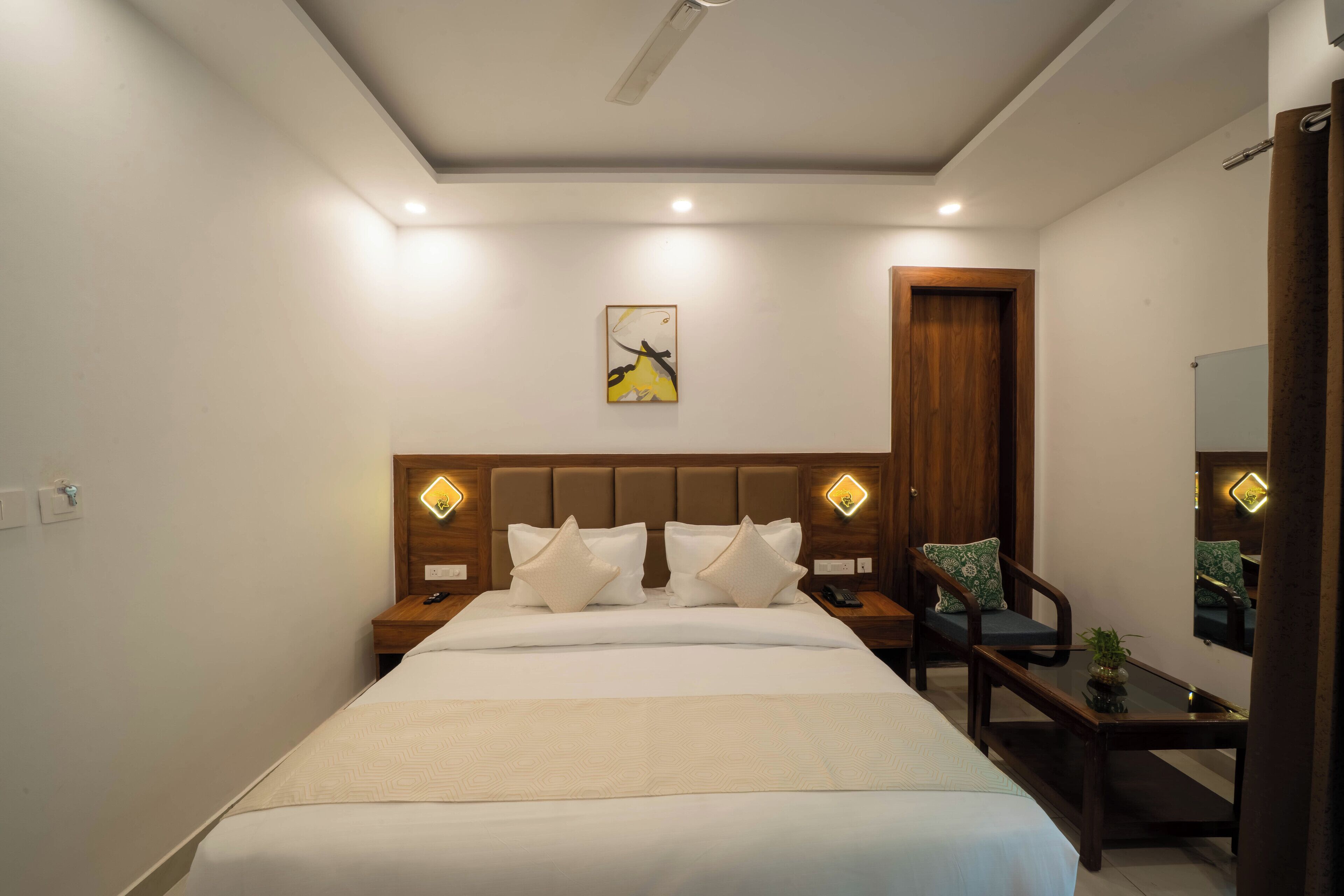 Une chambre d’hôtel avec un grand lit, deux tables de chevet, une tête de lit en bois, une commode, une chaise, une petite table avec une plante, un miroir et un tableau au mur.